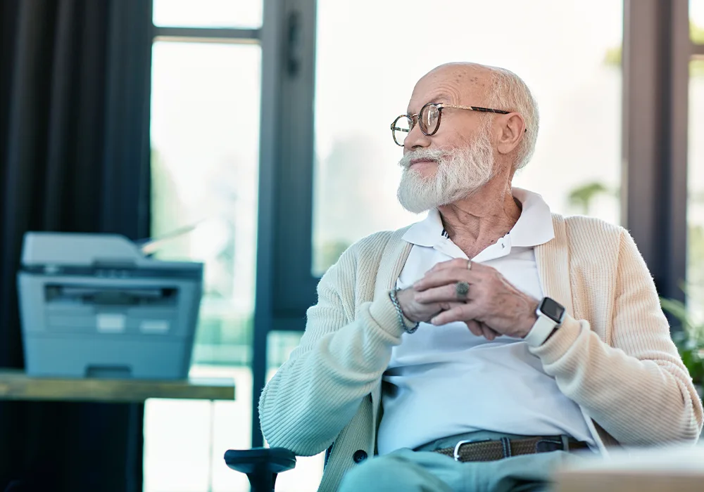 A thoughtful senior man with a white beard and glasses, wearing a light sweater, sitting by a window.